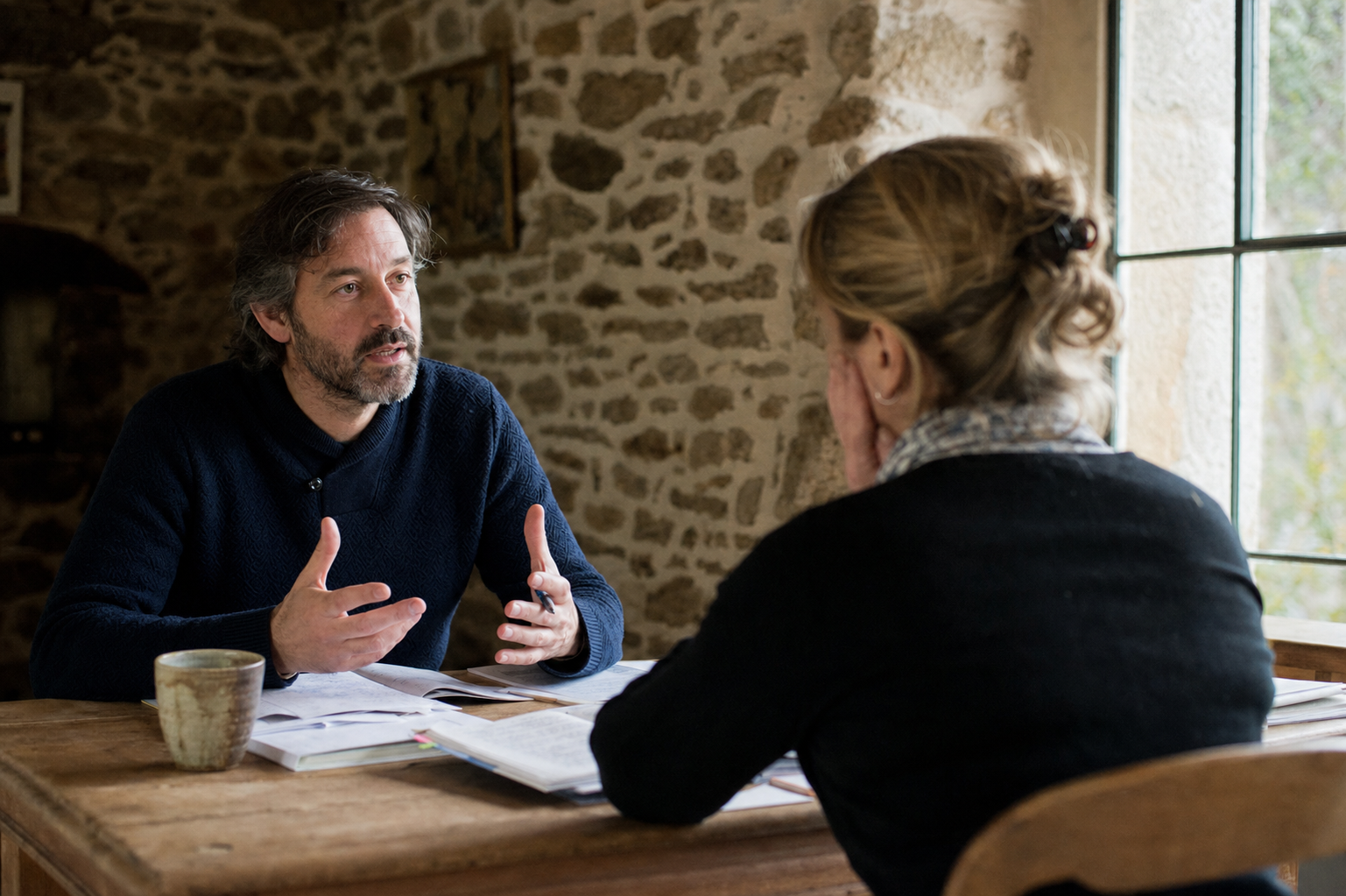 Two adults sit across a wooden table with papers and a cup as one speaks near a window.