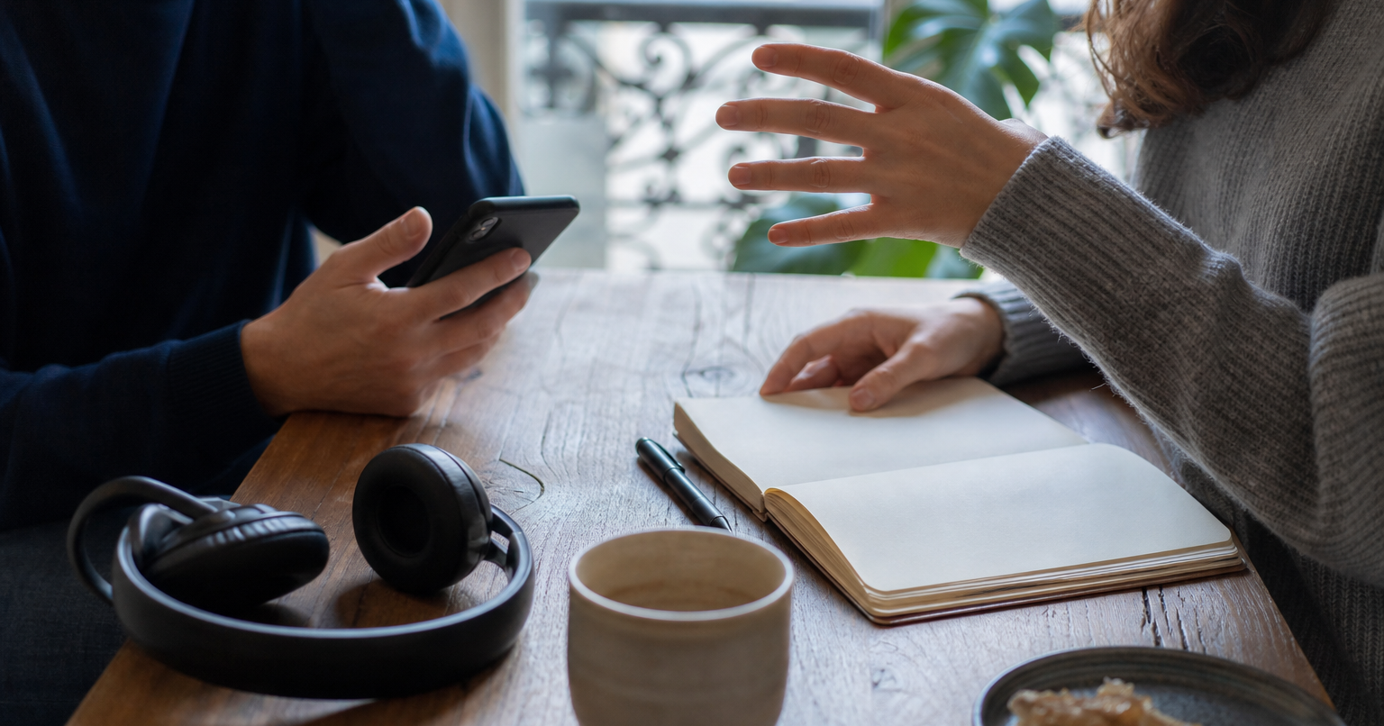 Two adults gesture across a wooden table with coffee, headphones, a notebook, pen, and closed unmarked laptop nearby.