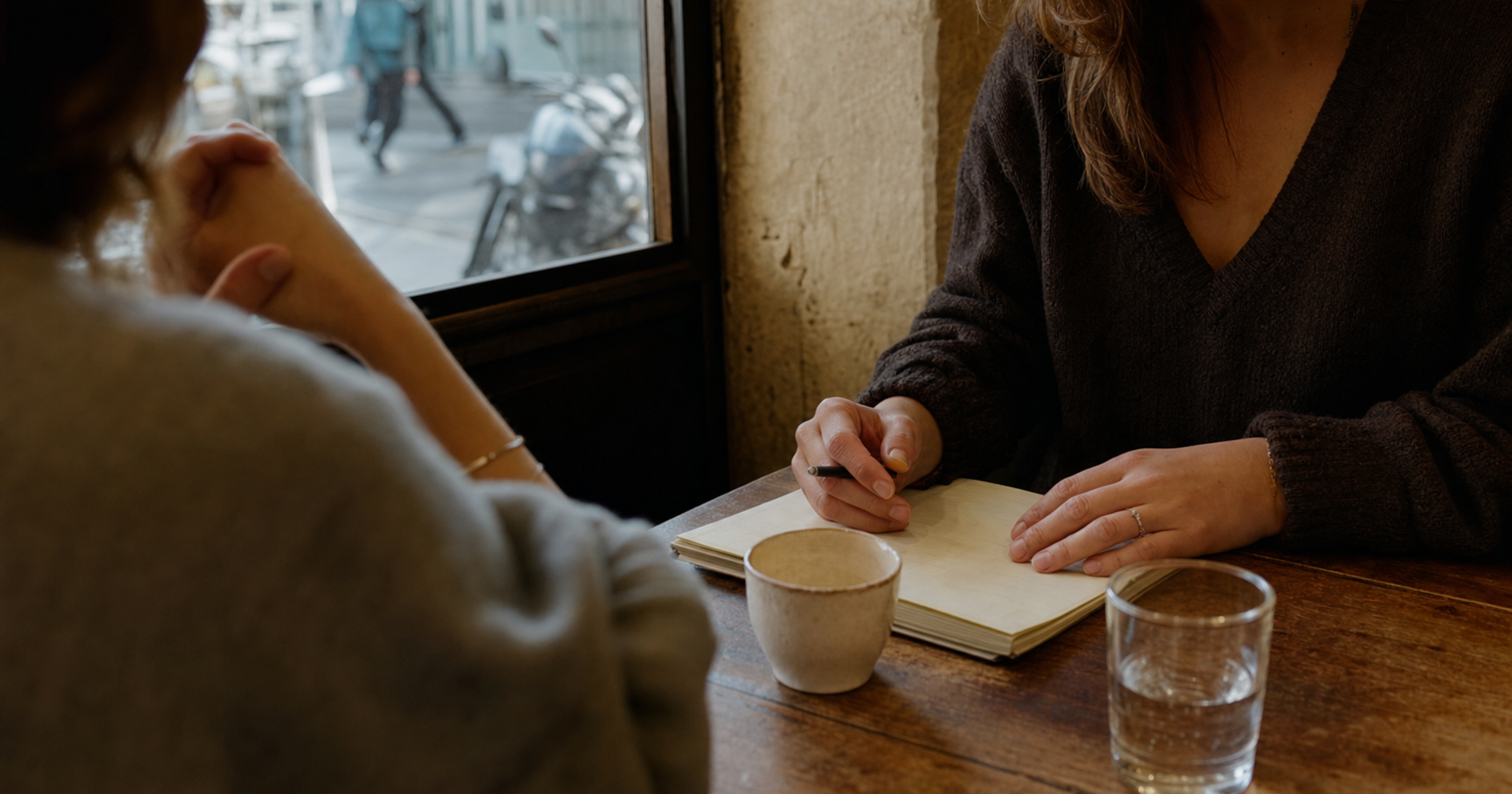 Two adults sit at a cafe table with a notebook, pen, coffee cup, and window view during conversation.