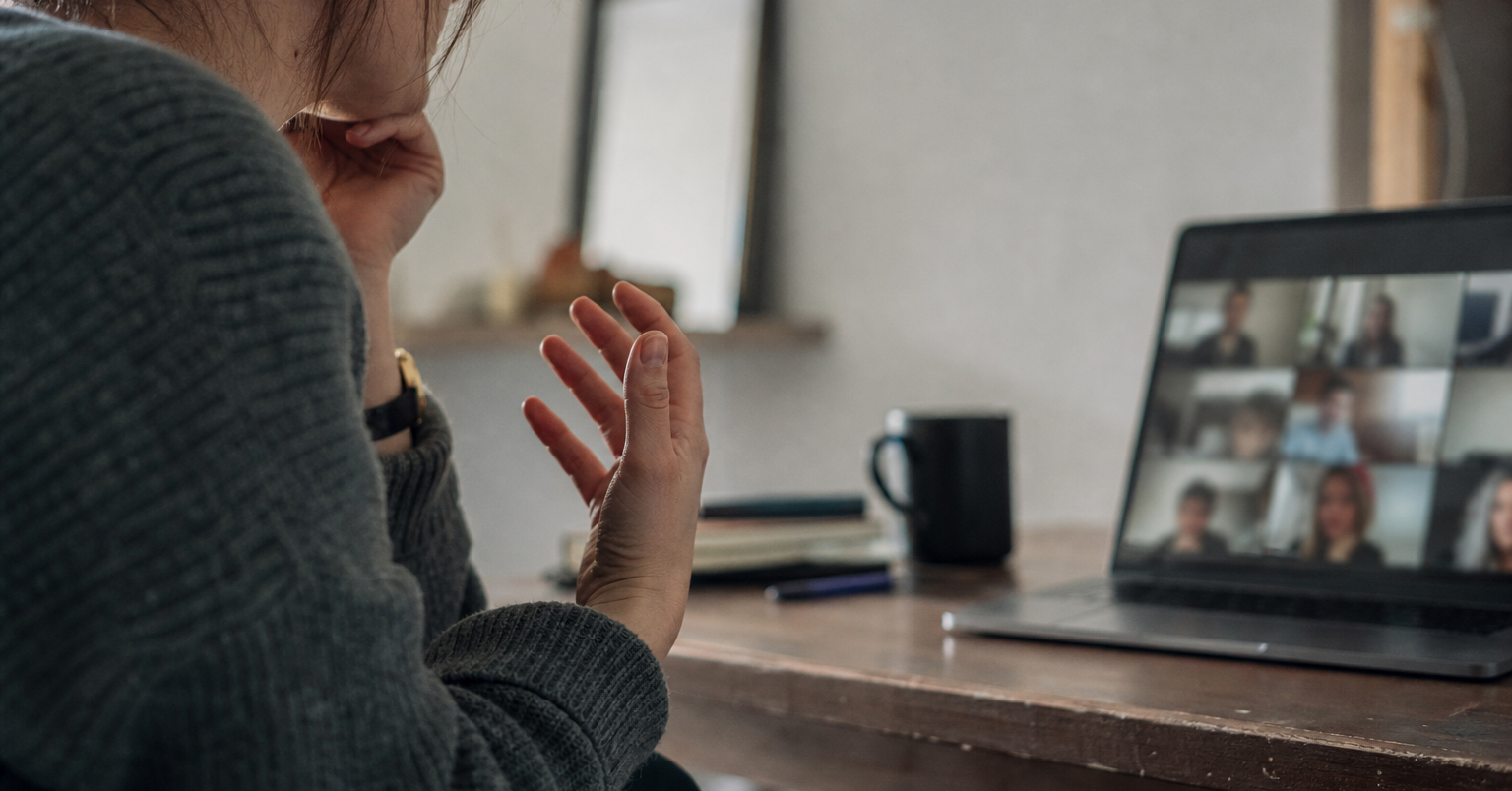 A person in profile gestures beside their mouth while attending a blurred online group class on a laptop.
