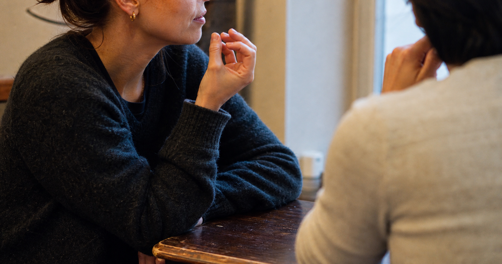 Two people sit close at a table, one speaking in profile with a raised hand while the listener faces away.